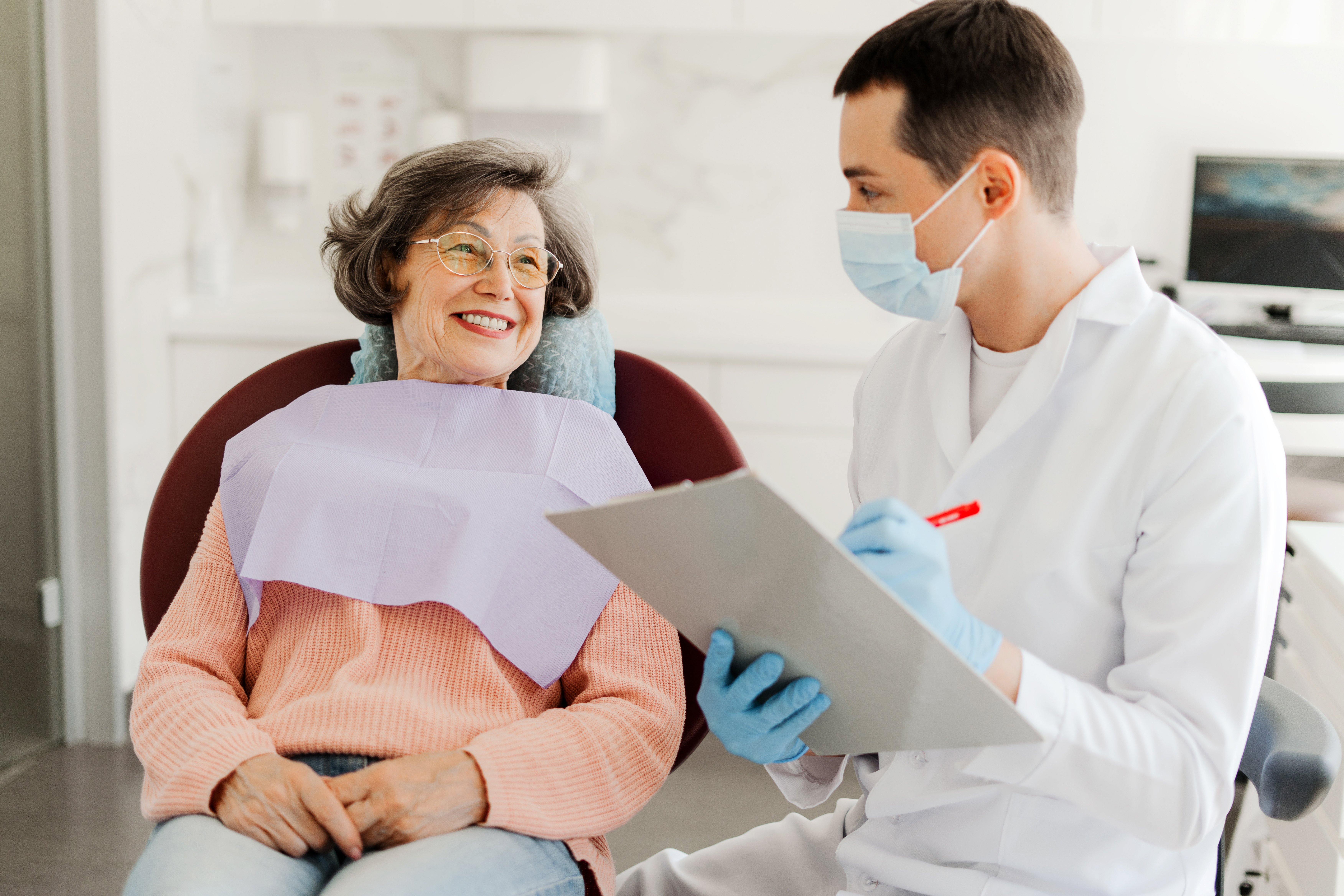 An older woman smiling in a dental chair while her dentist reviews a treatment plan on a clipboard.