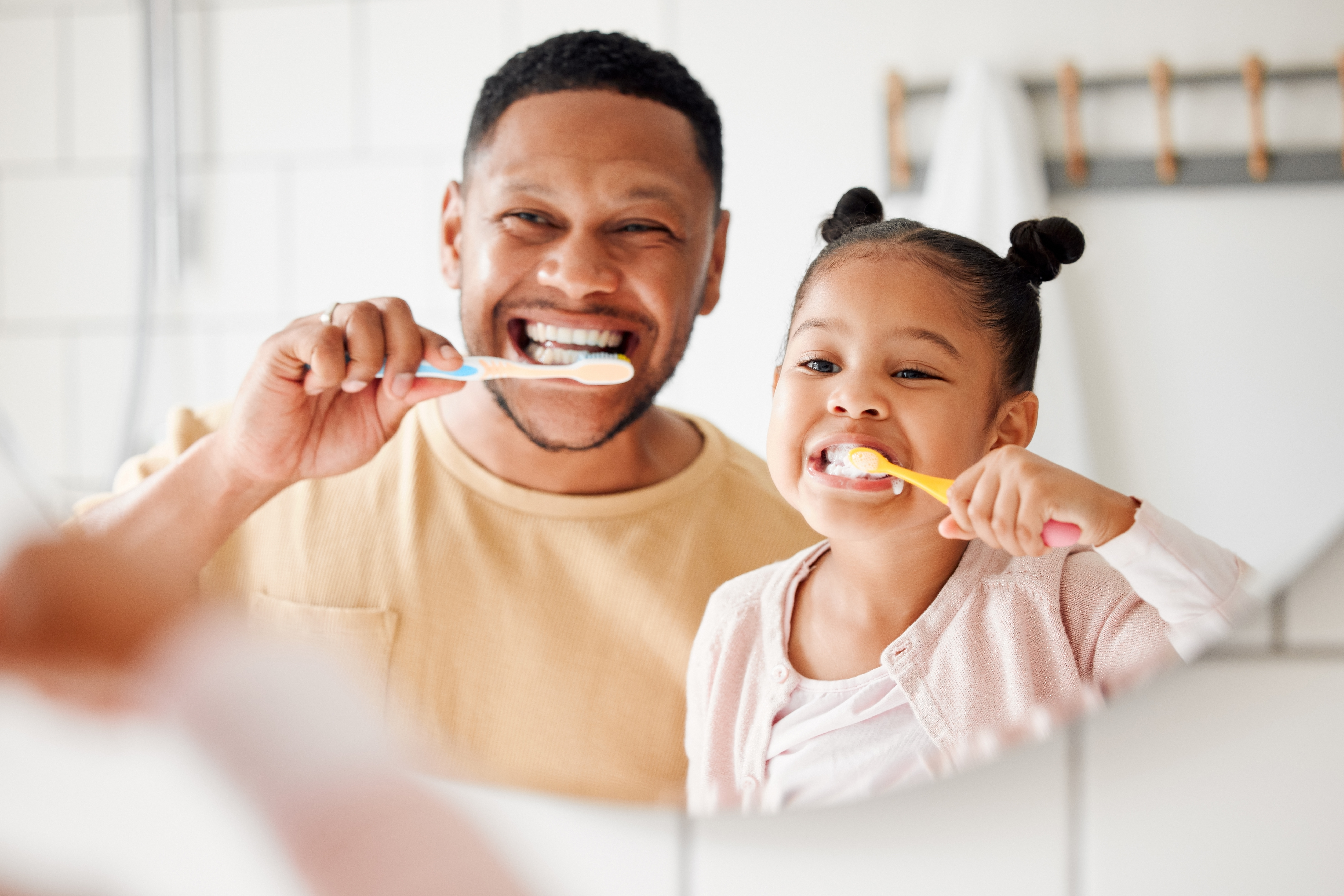 Father and daughter smiling as they brush their teeth looking at their reflection in the mirror.