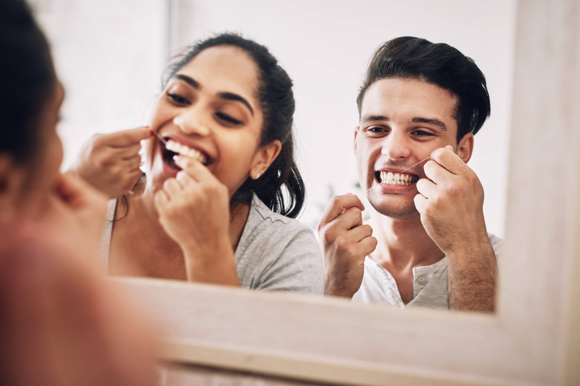 A couple flossing their teeth together while smiling in the bathroom mirror.